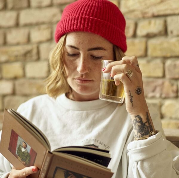 Frau mit weißem Pullover, roter Mütze, kurzen blonden Haaren, liest Buch und hält durchsichtiges Glas mit Bio Tee.