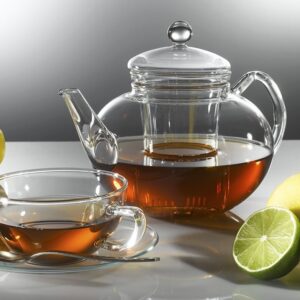 Teekanne mit Deckel und gläserne Tasse gefüllt mit Bio Tee auf Untertasse mit Teelöffel. Rechts eine halbe Limette.