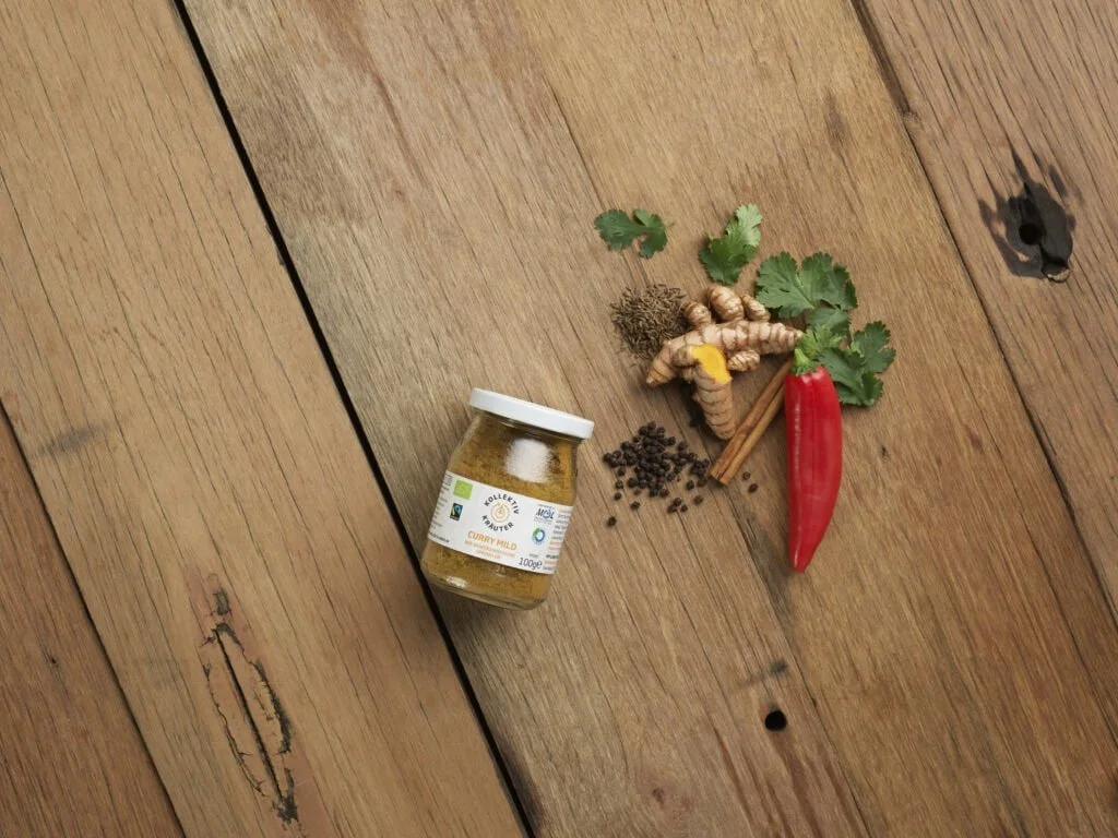 Bio Gewürz Curry Mild im Mehrwegglas, auf hölzernen Hintergrund mit Pfefferkörner, Zimtstange, Chilischote, Ingwar, Petersilie und Kümmel.