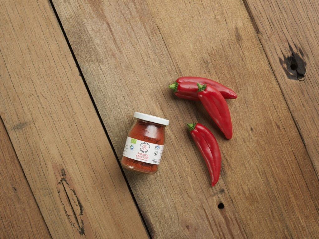 Bio Gewürz edelsüsse Paprika im Mehrwegglas, auf hölzernen Hintergrund mit drei roten Paprikaschoten.