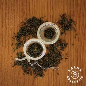 Fairtrade Bio Darjeeling verstreut auf hölzernem Hintergrund, mittig ein halbgefülltes Teeei offen.