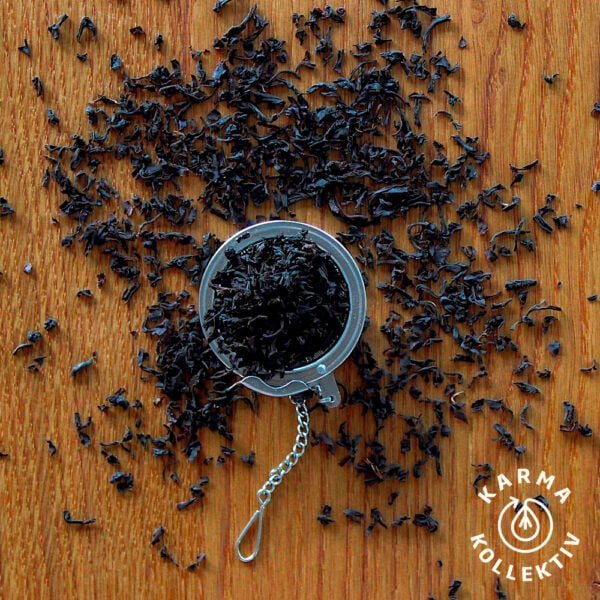 Fairtrade Bio Schwarzer Tee verstreut auf hölzernem Hintergrund, mittig ein halbgefülltes Teeei offen.